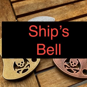 Op de afbeelding: Drie metalen sleutelhangers met een schedel en gekruiste beenderen ontwerp. De sleutelhangers zijn in zilver, goud en koper. De tekst "Ship's Bell" wordt weergegeven op een zwarte achtergrond.