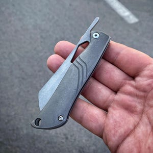 May include: A folding pocket knife with a matte grey metal handle featuring three curved engraved lines. The knife has a prominent flipper tab, a visible pivot screw, and a lanyard hole at the base. The blade is partially retracted into the handle.