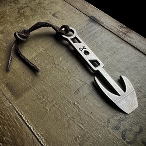 May include: A silver metal bottle opener with a hook design and a leather cord. The opener has the letters "X" and "O" etched into the metal. The leather cord is dark brown and knotted.