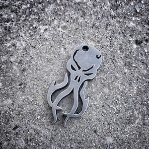 Puede incluir: Un llavero de metal plateado con forma de calavera con tentáculos. El llavero es un diseño detallado con una cara de calavera y tentáculos.