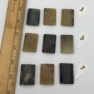 May include: Nine rectangular, polished stones in shades of gray, brown, and tan are arranged on a white surface. A wooden ruler is positioned along the left side. Three small white paper triangles are labeled with the numbers 1, 2, and 3.