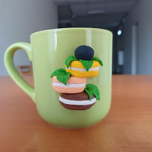 Può includere: Una tazza in ceramica verde chiaro decorata con tre colorati macarons e un mirtillo. I macarons sono gialli, pesca e marroni, con ripieno bianco e dettagli di foglie verdi. La tazza ha un manico curvo.