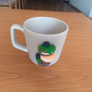 Può includere: Una tazza in ceramica color crema con un manico bianco. La tazza è decorata con una pila di tre colorati macarons, ognuno con un colore diverso e sormontato da un mirtillo e foglie verdi.