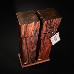 Op de afbeelding: Twee houten zout- en peperstrooiers met een natuurlijk, rustiek ontwerp. De strooiers zijn gemaakt van donkerbruin hout en staan op een kleine houten basis. Er is een label aan een van de strooiers bevestigd.