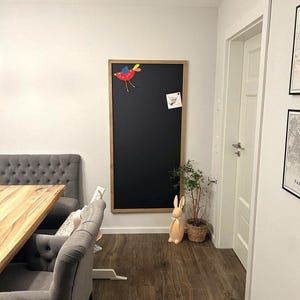 May include: A large rectangular chalkboard with a light wood frame mounted on a white wall. A colorful red and blue bird cutout is attached to the top left, and a small square white note is pinned to the upper right side of the black surface.