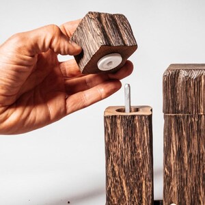 May include: A set of two wooden salt and pepper shakers with a rustic, natural wood finish. The shakers have a square shape and are made of dark wood with a rough texture. The top of the shaker is removable, revealing a white ceramic grinder.