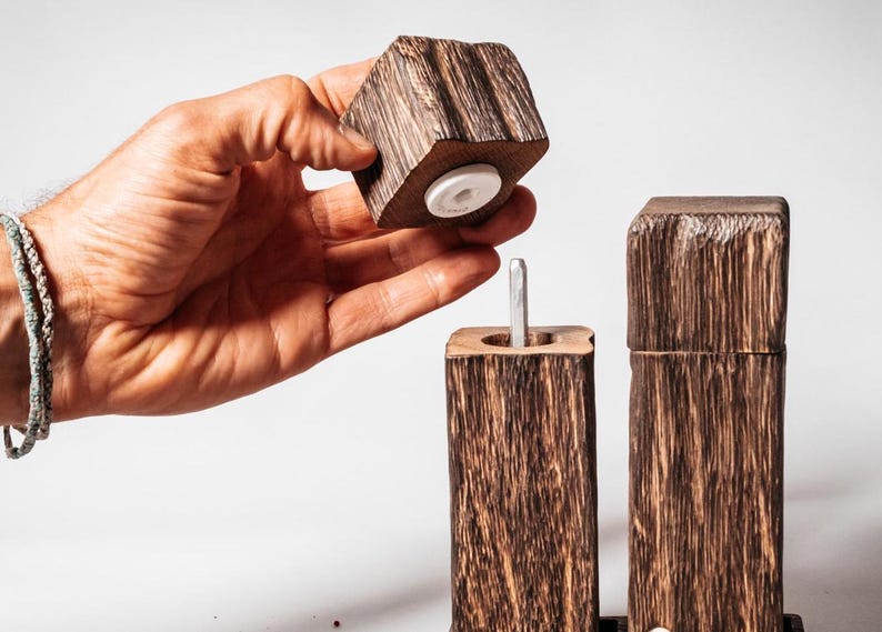 May include: A set of two wooden salt and pepper shakers with a rustic, natural wood finish. The shakers have a square shape and are made of dark wood with a rough texture. The top of the shaker is removable, revealing a white ceramic grinder.