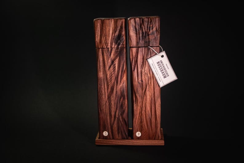 Op de afbeelding: Een paar houten zout- en pepermolens met een getextureerde, donkerbruine afwerking. De rechthoekige molens staan op een kleine houten basis. Een label met Duitse tekst hangt eraan.
