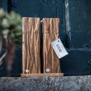 May include: Two wooden salt and pepper grinders with a textured, bark-like appearance. The grinders are light brown and sit on a wooden base. A tag with the word "EICHE" is attached to one of the grinders.