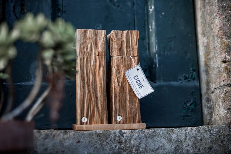 May include: Two wooden salt and pepper grinders with a textured, bark-like appearance. The grinders are light brown and sit on a wooden base. A tag with the word "EICHE" is attached to one of the grinders.