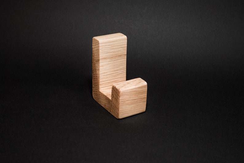 May include: A wooden wall hook in a natural oak finish. The hook has a simple, L-shaped design with a rectangular base and a shorter, square-shaped hook. The wood grain is visible, adding texture. The hook is set against a dark background.