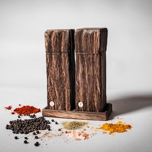May include: Two wooden salt and pepper shakers with a dark brown finish, sitting on a wooden base. The shakers are labeled "S" and "P". The shakers are surrounded by various spices, including black peppercorns, red pepper flakes, turmeric, and dried herbs.