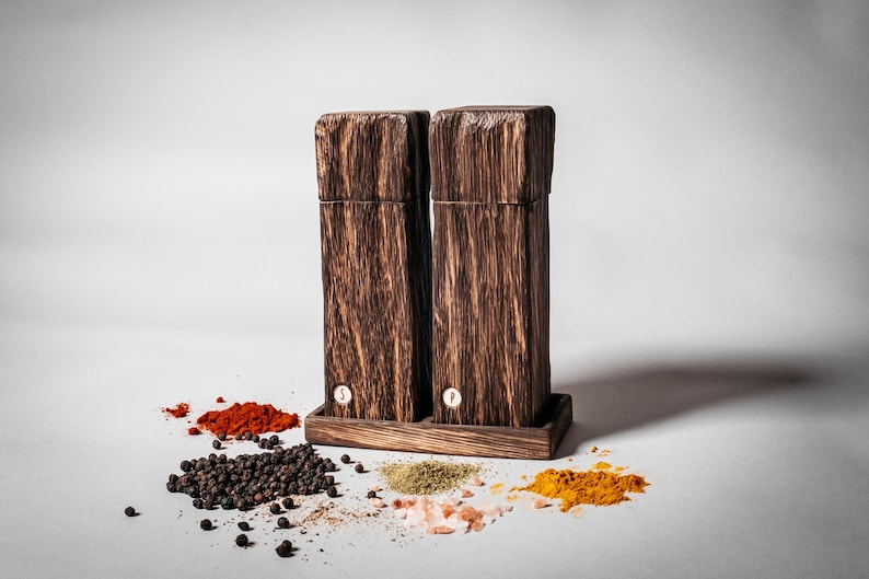 May include: Two wooden salt and pepper shakers with a dark brown finish, sitting on a wooden base. The shakers are labeled "S" and "P". The shakers are surrounded by various spices, including black peppercorns, red pepper flakes, turmeric, and dried herbs.