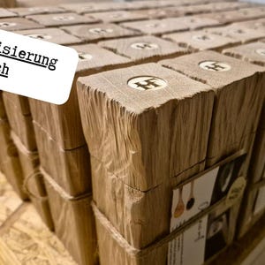 May include: A collection of wooden blocks, each with a carved letter and wrapped in paper. The blocks are arranged in rows, with the text "Individualisierung m&ouml;glich" in a white box. Silhouettes of trees are in the corner.