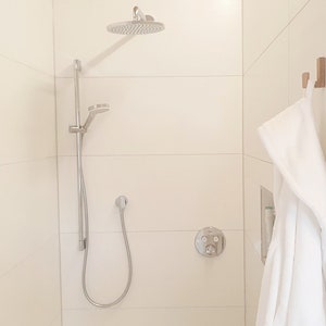 May include: A modern shower with a silver shower head and handheld sprayer. The shower is in a white tiled enclosure. A white robe hangs on a wooden hook. The shower fixtures are silver.