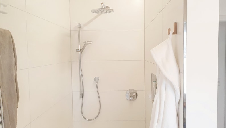 May include: A modern shower with a silver shower head and handheld sprayer. The shower is in a white tiled enclosure. A white robe hangs on a wooden hook. The shower fixtures are silver.