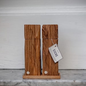 May include: Two wooden salt and pepper grinders with a natural wood grain. The rectangular grinders have a square top and sit on a small wooden tray. A tag reading "EICHE" hangs from one. The grinders are labelled "S" and "P".