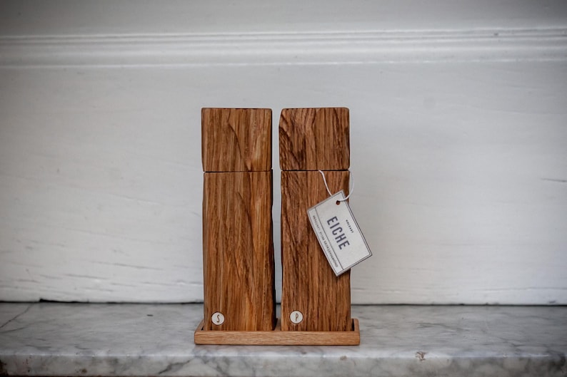 May include: Two wooden salt and pepper grinders with a natural wood grain. The rectangular grinders have a square top and sit on a small wooden tray. A tag reading "EICHE" hangs from one. The grinders are labelled "S" and "P".
