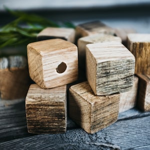 Può includere: Un insieme di piccoli cubi di legno naturale con venature visibili e un aspetto rustico. Un cubo ha un foro traforato. Rami verdi sono visibili sullo sfondo, suggerendo un tema naturale. I cubi sono disposti su una superficie di legno invecchiato.