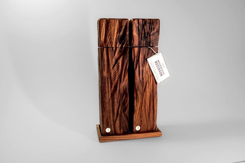 Op de afbeelding: Een hoge, rechthoekige houten sculptuur met een textuur die op schors lijkt. Het donkerbruine hout is gemonteerd op een kleine, rechthoekige basis. Een label is bevestigd met de tekst "SCHWEIZERISCHER NUSSBAUM".