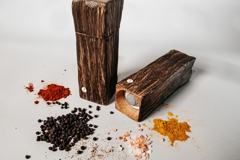 May include: Two wooden spice grinders with a rustic finish. The grinders are filled with different spices, including black peppercorns, paprika, turmeric, and pink Himalayan salt.