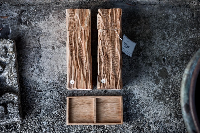 May include: Two wooden salt and pepper grinders with a textured, bark-like design, and a small wooden tray. The grinders have a label that reads "Eiche". One grinder is marked with an "S" and the other with a "P".