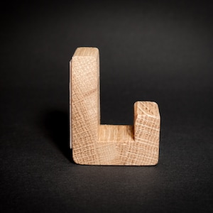 May include: A wooden letter "L" stands upright against a black background. The letter is constructed from light-colored wood, showcasing the natural grain. The design is simple and modern, suitable for home decor or craft projects.