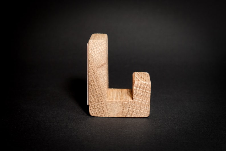 May include: A wooden letter "L" stands upright against a black background. The letter is constructed from light-colored wood, showcasing the natural grain. The design is simple and modern, suitable for home decor or craft projects.