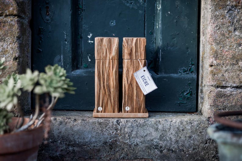 May include: Two wooden salt and pepper grinders with a natural wood grain finish. The grinders are rectangular and sit on a wooden base. A tag that says "EICHE" hangs from one of the grinders. The background is a dark teal door.