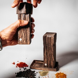 May include: Two wooden salt and pepper shakers with a dark brown finish. The shakers are made of wood and have a simple design. The shakers are being used to season food, with a white powder coming out of one of the shakers.