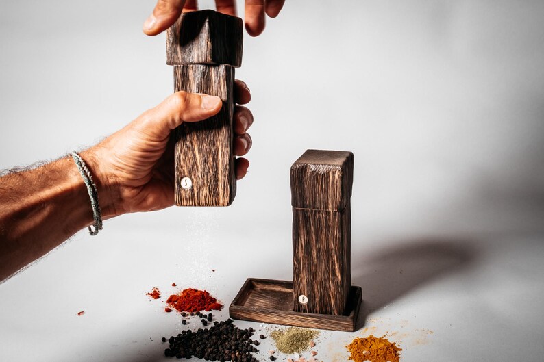 May include: Two wooden salt and pepper shakers with a dark brown finish. The shakers are made of wood and have a simple design. The shakers are being used to season food, with a white powder coming out of one of the shakers.