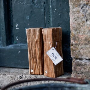 May include: Two wooden rectangular boxes with a natural wood grain finish. One box has a tag that reads "EICHE". The boxes are leaning against a dark green door and a stone wall.