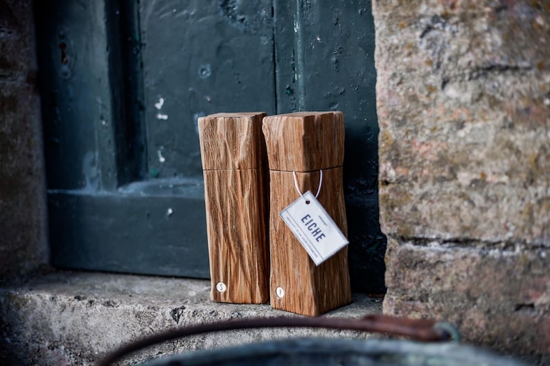May include: Two wooden rectangular boxes with a natural wood grain finish. One box has a tag that reads "EICHE". The boxes are leaning against a dark green door and a stone wall.