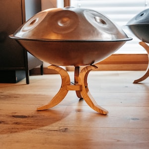 May include: Two handpan drums on wooden stands. The handpans are both round and have a metallic finish. The stand is made of light brown wood and has a curved design.