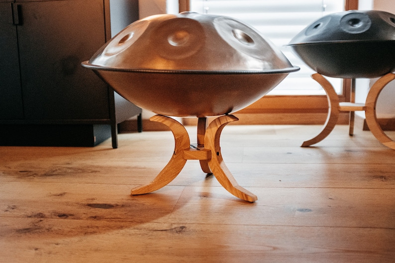May include: Two handpan drums on wooden stands. The handpans are both round and have a metallic finish. The stand is made of light brown wood and has a curved design.