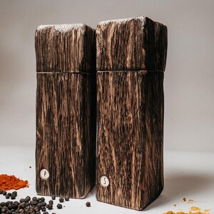 May include: A set of two wooden salt and pepper shakers. The shakers are made from dark wood with a natural grain pattern. The shakers are labeled "P" and "S" for pepper and salt.