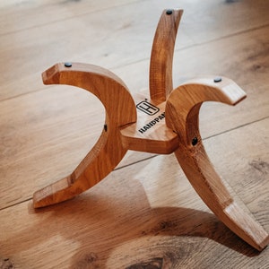 May include: A wooden stand with three curved legs and a flat top. The stand is made of light brown wood and has a black label on the top that says "HANDPAN".