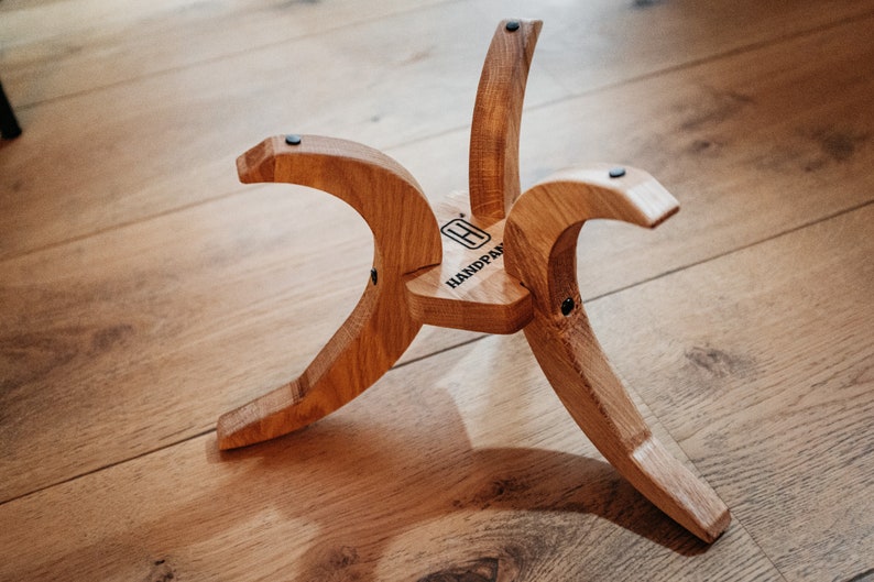 May include: A wooden stand with three curved legs and a flat top. The stand is made of light brown wood and has a black label on the top that says "HANDPAN".