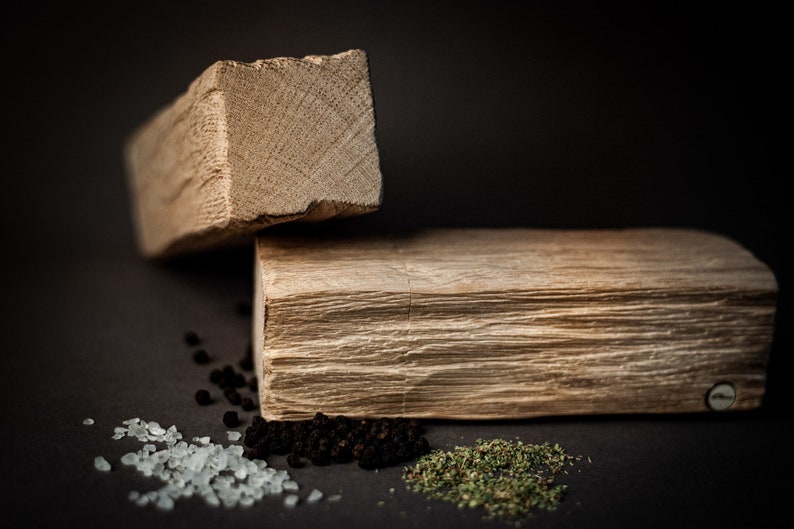 May include: Two wooden blocks, one stacked on the other, against a dark backdrop. Small piles of salt, peppercorns, and dried herbs are scattered around the blocks. The wood has a natural, textured appearance.