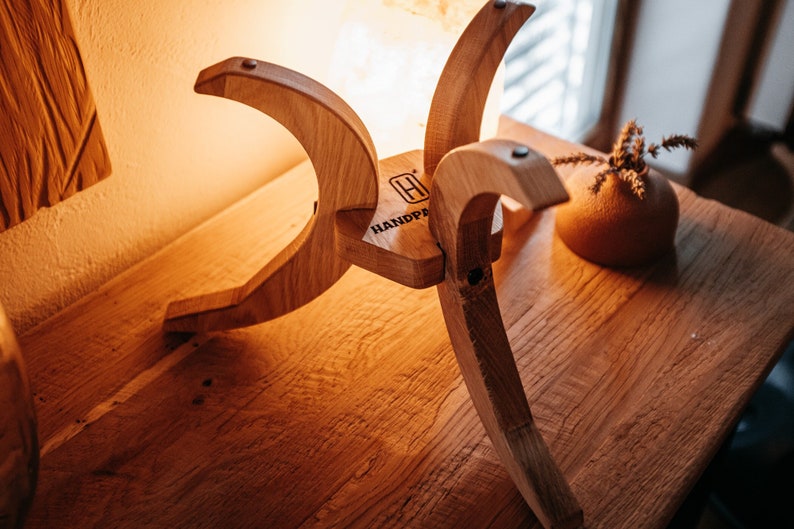 May include: A wooden lamp stand with a unique design, featuring four curved legs and a flat top. The stand is made of natural wood and has a rustic look. The stand is labelled "HANDMADE" on the top.