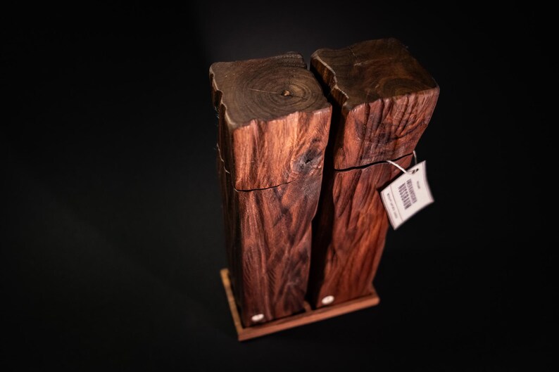 Op de afbeelding: Twee houten zout- en peperstrooiers met een natuurlijk, rustiek ontwerp. De strooiers zijn gemaakt van donkerbruin hout en staan op een kleine houten basis. Er is een label aan een van de strooiers bevestigd.