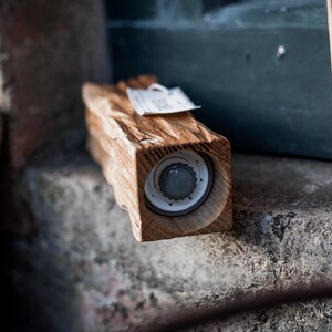 May include: A wooden salt or pepper grinder with a natural, rustic finish. The grinder is rectangular with a visible grinding mechanism and a small tag attached. The wood grain is prominent, and the overall aesthetic is handcrafted.