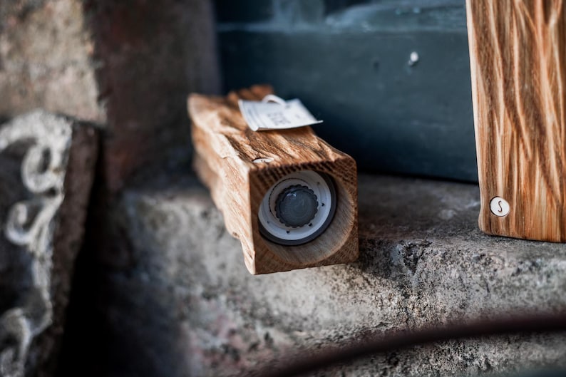 May include: A wooden salt or pepper grinder with a natural, rustic finish. The grinder is rectangular with a visible grinding mechanism and a small tag attached. The wood grain is prominent, and the overall aesthetic is handcrafted.