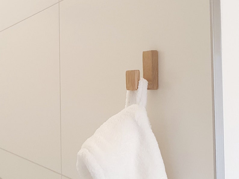 May include: Two wooden wall hooks with a white towel hanging from one. The hooks are rectangular and made of light-colored wood. The towel is fluffy and white, contrasting with the wood and the neutral wall.