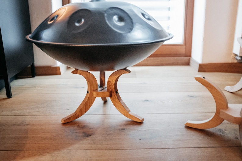 May include: A dark grey steel handpan sits on a wooden stand with three legs. The stand is made of light brown wood and has a curved design.