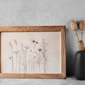 Könnte beinhalten: Eine gerahmte Schwarzweiß-Zeichnung von Wildblumen in einem Holzrahmen. Der Rahmen steht auf einer grauen Betonfläche. Rechts vom Rahmen steht eine schwarze Vase mit getrockneten Mohnkapseln.