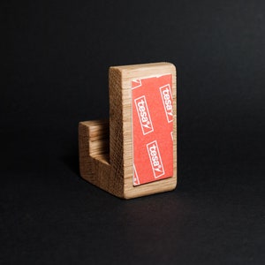 May include: A wooden wall hook with a right-angle design. The hook is made of light-colored wood and features a red and white adhesive strip on the back, with the word "tesa" printed on it. The hook is set against a black background.