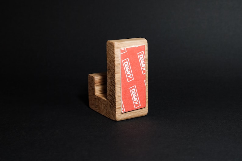 May include: A wooden wall hook with a right-angle design. The hook is made of light-colored wood and features a red and white adhesive strip on the back, with the word "tesa" printed on it. The hook is set against a black background.