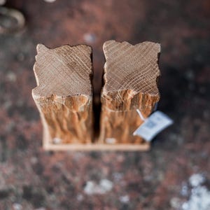 May include: Two wooden bookends with a natural, rough-hewn texture. The bookends are made of light brown wood and have a rectangular base. The top edges have a unique, uneven shape. A small tag hangs from one of the bookends.