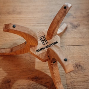 May include: A wooden handpan stand with a hexagonal base and four legs. The stand is engraved with the text "HANDPANSTAND" and a stylised "H".